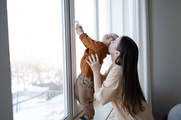 A mother and a small child stand at a large white window and look out into the street. Safety for small children in the house. - obrazy, fototapety, plakaty