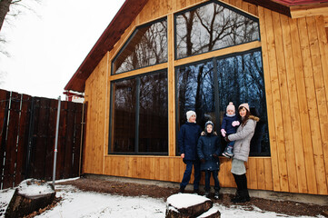 Attractive mother with her kids in winter day stand near wooden house with large window. - obrazy, fototapety, plakaty