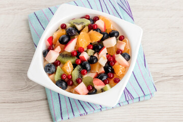 Fruit fresh salad for a breakfast snack of apples, tangerines, kiwis, lingonberries, blueberries in a white bowl. Healthy eating concept.