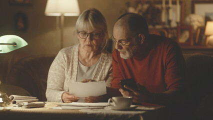 Elderly husband and wife analyzing utility bills and making calculations on smartphone while sitting near table in living room at home