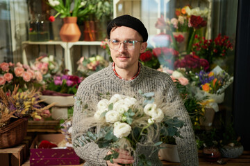 Cute smiling bearded man with bouquet of roses in flower shop. Good looking male florist wearing sweater, beanie and eyeglasses holding flower bouquet. Small business concept. High quality photo