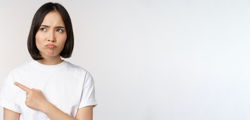 Angry and upset young asian woman pointing left, looking with regret or jealousy, feel unfair,...