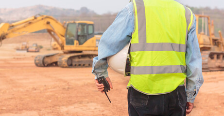 An engineer or worker wearing a life jacket holds a radio transmitter and a white helmet in his hands. for safe working on excavator background