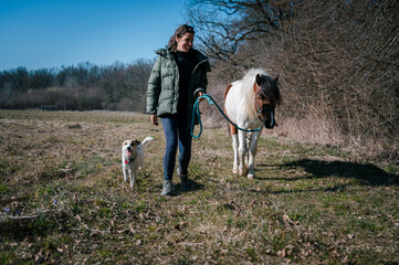 No make-up young woman with a big smile walking in nature together with a beautiful pony and a dog....