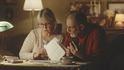 Preoccupied senior husband and wife analyzing papers and making calculations while checking utility bills in dim living room at home