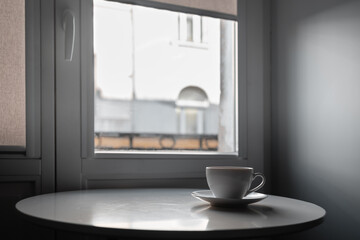 A lonely white cup in the morning on a table by the window against the wall of a neighboring house in a poor apartment. The concept of loneliness, asceticism and minimalism - obrazy, fototapety, plakaty