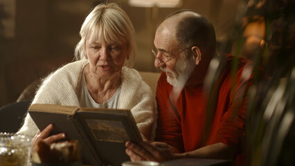 Cheerful elderly wife speaking with husband and laughing while looking at pictures in photo album while sitting on couch in living room at home