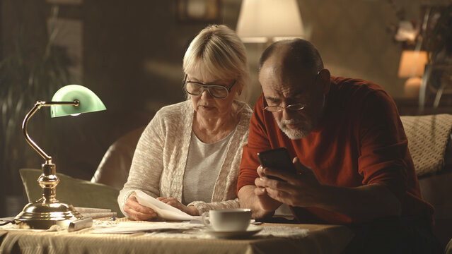 Preoccupied senior husband and wife analyzing papers and making calculations while checking utility bills in dim living room at home