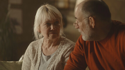 Elderly husband and wife speaking with each other during quarrel on couch in living room at home