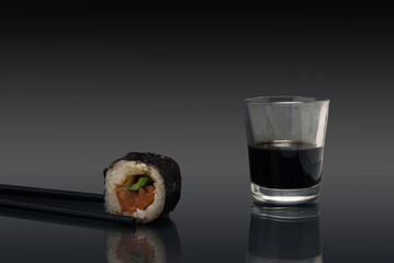 Sushi minimal concept. Chopsticks, one piece of rice, salomon and cucumber roll in nori dried edible seaweed beside soy sos glass. Awesome vibe with black background
