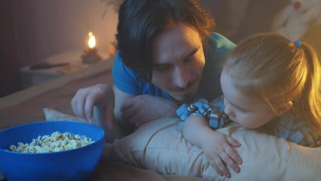 Dad and daughter watch TV, cartoon, evening. Lying on bed, resting, spending time, lazy leisure. Family relationships, love, man and child, Father's Day. eat popcorn and play