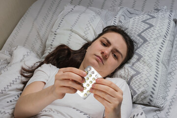 A tired sleepy woman lies in bed, holding her head with her hand, pills in her hands.