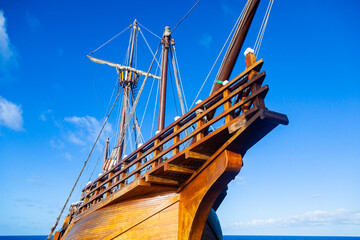An old wooden caravel ship with folded sails against a blue sky with white clouds.