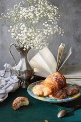 Still life with freshly baked croissants on a plate, an open book, white gypsophila flowers in vintage silver vase. Weekend breakfast concept. 