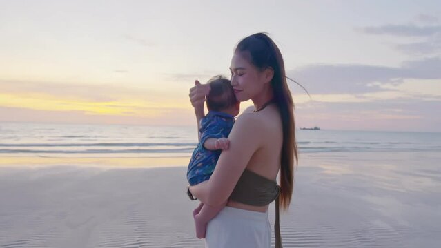 Happy mother with baby boy walks by ocean on the beach in summer. Both are having fun time and cheerful together. Motherhood love and care for her child. An amazing view retreats them a lot.