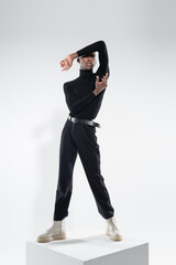 full length of young african american man in totally black outfit standing on white cube while posing on grey