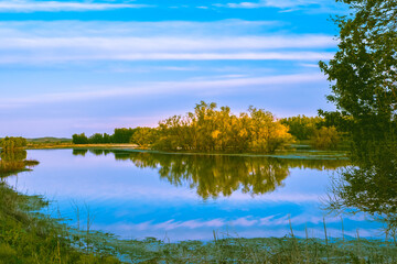 Fototapeta na wymiar Picturesque view of Midwestern lake with trees in background at sunset in fall; trees and blue sky with light clouds reflect in calm water