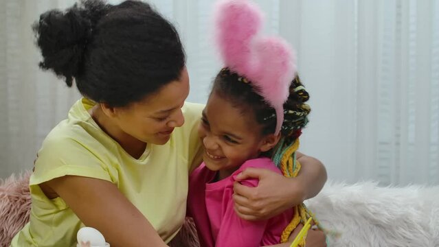 Mom and daughter with funny bunny ears happily hugging and smile. African American woman and little girl are sitting in decorated room at home. Happy easter. Slow motion. Close up.