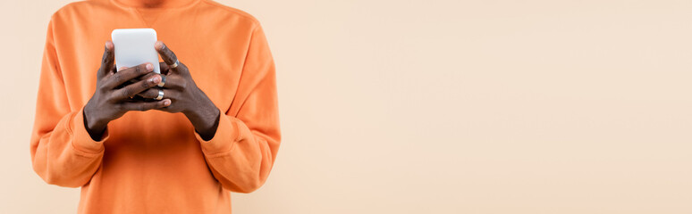 cropped view of african american man in orange sweatshirt using smartphone isolated on beige, banner
