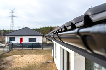 A metal, black gutter on a roof covered with ceramic tiles, visible house in the background.