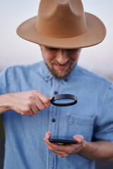 Male wearing jeans shirt and hat looking with loupe at smartphone as detective, investigator or...