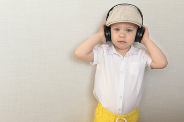 A little boy in a white shirt, yellow shorts and a retro cap listens to music through large headphones.