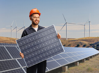 Worker in a uniform holding a solar panel on a solar and wind turbine farm