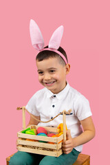 Cheerful boy sitting on a chair wearing rabbit ears holding basket with eggs on his knees posing on pink background.