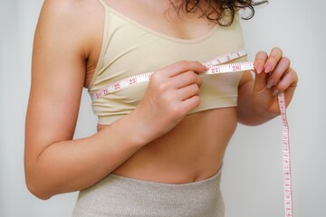 Cropped view of slim woman measuring breasts with tape measure at home, close up. Unrecognizable European woman checks the result of a weight loss diet or liposuction indoors. Healthy lifestyle.