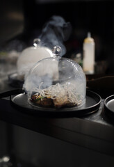 Plato de comida fusión emplatado con cubre platos de cristal y humo de aroma saliendo del plato