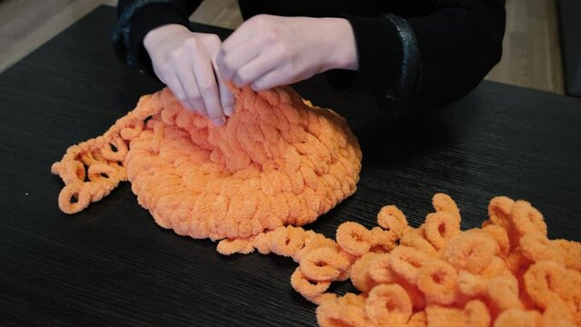 The girl knits with her hands a balaclava of orange color with the help of Alize Puffy threads. Half the work is done.