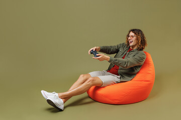 Full size body length gambling young brunet curly man 20s wears khaki shirt sit in bag chair hold...