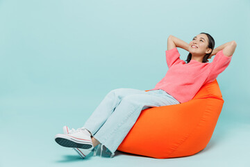 Full body young smiling minded fun woman of Asian ethnicity 20s wear pink sweater sit in bag chair hold hands behind neck isolated on pastel plain light blue color background People lifestyle concept. - Powered by Adobe