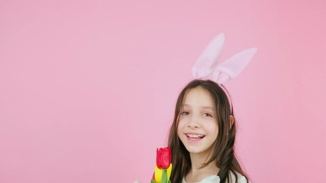 Croppde video of a funny girl wearing bunny ears hiding face with flowers and then takes them off the face on pink background.
