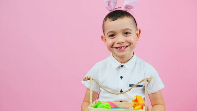 Little cute boy wearing rabbit ears sitting on a chair holding basket with Easter eggs on pink background and copy space.