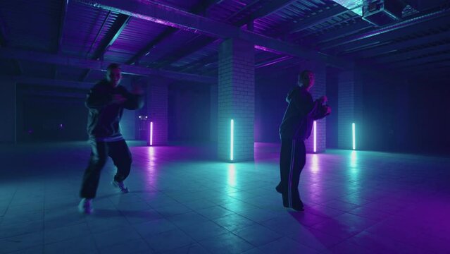 Two hip-hop dancers in neon light dance together in blue and purple colors. Women enjoying hip hop moves in dark studio with smoke and lighting.