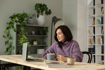 a cute brunette girl in a lilac sweater is sitting at a table with a laptop