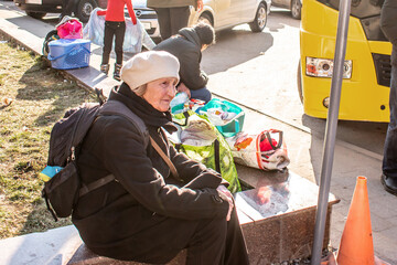Sad elderly woman with a Ukrainian flag in a backpack near the evacuation buses. Grandmother with...