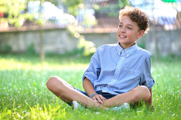 Naklejka premium Young happy child boy relaxing on green grass in summer park. Positive kid enjoying summertime outdoors. Child wellbeing concept