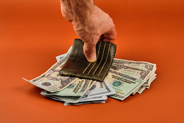 A man's hand crushing a wallet full of dollars on an orange table.