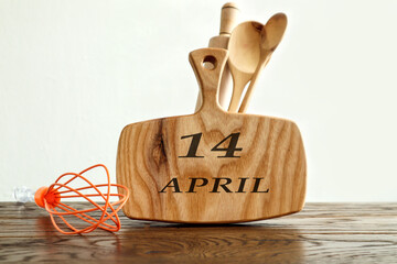 April 14 calendar: on a kitchen wooden board the name of the month April in English, the number 14, next to various kitchen utensils, a gray background