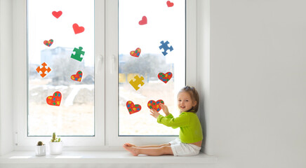 A cute little happy girl decorated the window in her room with hearts and puzzles as a sign of...