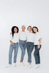 Fototapeta premium Full length of positive interracial women hugging on grey background.