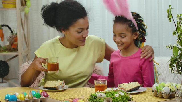 Mom and daughter with funny bunny ears are drinking tea with Easter cake. African American woman and little girl are sitting at table in festively decorated room at home. Slow motion. Close up.