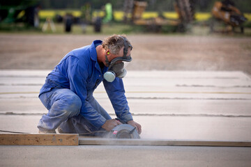 A man wearing a face mask cutting through concrete with a circular saw outdoors in summer