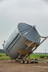 A steel grain bin tipped on a grain bin mover in a summer countryside landscape