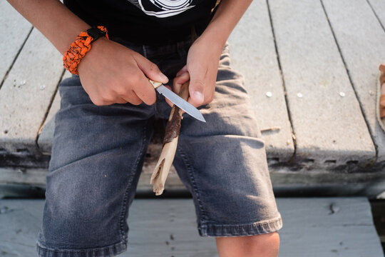 Boy sitting on the edge of a dock on a lake whittling a piece of wood