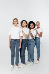 Fototapeta premium Full length of multiethnic women in jeans and white t-shirts looking at camera on grey background.