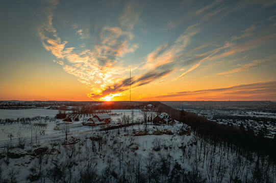 An aerial view of a beautiful vibrant golden-orange sunset above a red barn perched on a snowy forested hill during winter in Hamilton, Ontario, Canada.