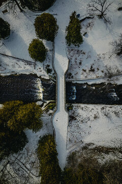 An aerial top-down view of green trees and a bridge over a flowing river with dark water on a snowy winter morning, in Hamilton, Ontario, Canada.